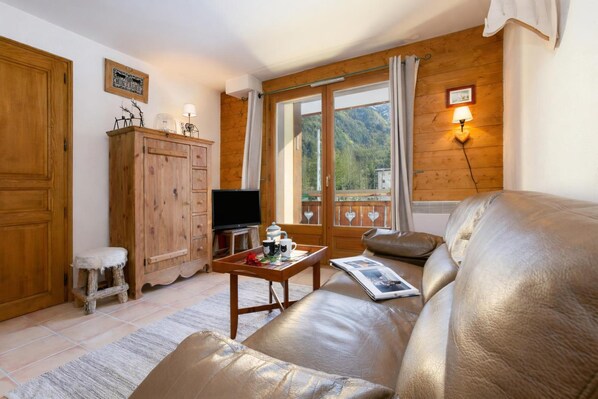 A cozy living room with a leather sofa, wooden cabinets, a television, and a balcony with a mountain view.