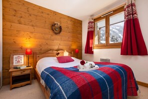 A cozy bedroom with a wooden headboard, a bed with a red and blue plaid blanket, two bedside lamps, a framed picture, and a window with red curtains. - Apt Capucins22 Mont Blanc View 1BR w/ Balcony Garage, Chamonix, France (Chamonix)