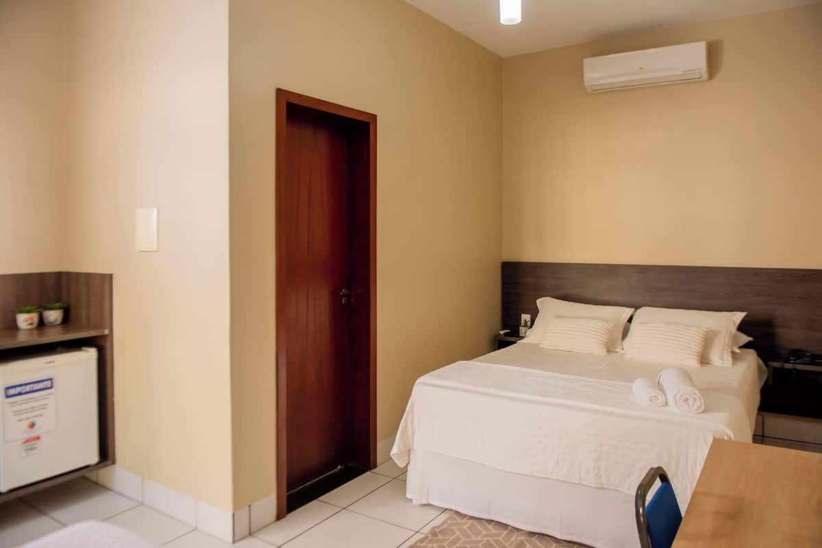 A hotel room with a bed, a desk, a chair, a wardrobe, and an air conditioning unit.