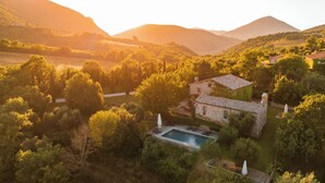 Un bâtiment rustique en pierre, doté d’une piscine, entouré d’une végétation luxuriante et avec des montagnes en toile de fond.