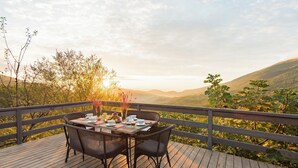 Une terrasse en bois avec une table dressée pour deux personnes, entourée de chaises, d’où l’on peut admirer un paysage montagneux avec un coucher de soleil en arrière-plan.