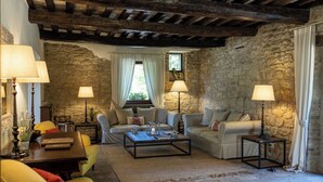Un salon rustique avec des murs en pierre, des poutres en bois et une cheminée en pierre. On y trouve deux canapés, une table basse et un fauteuil jaune. Des lampes et un vase rempli de fleurs sont posés sur la table d’appoint.