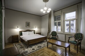 A bedroom with a wooden bed, two bedside lamps, a green armchair, a wooden table, and a large window with blinds. - Casa Gotica Residence (Brasov)