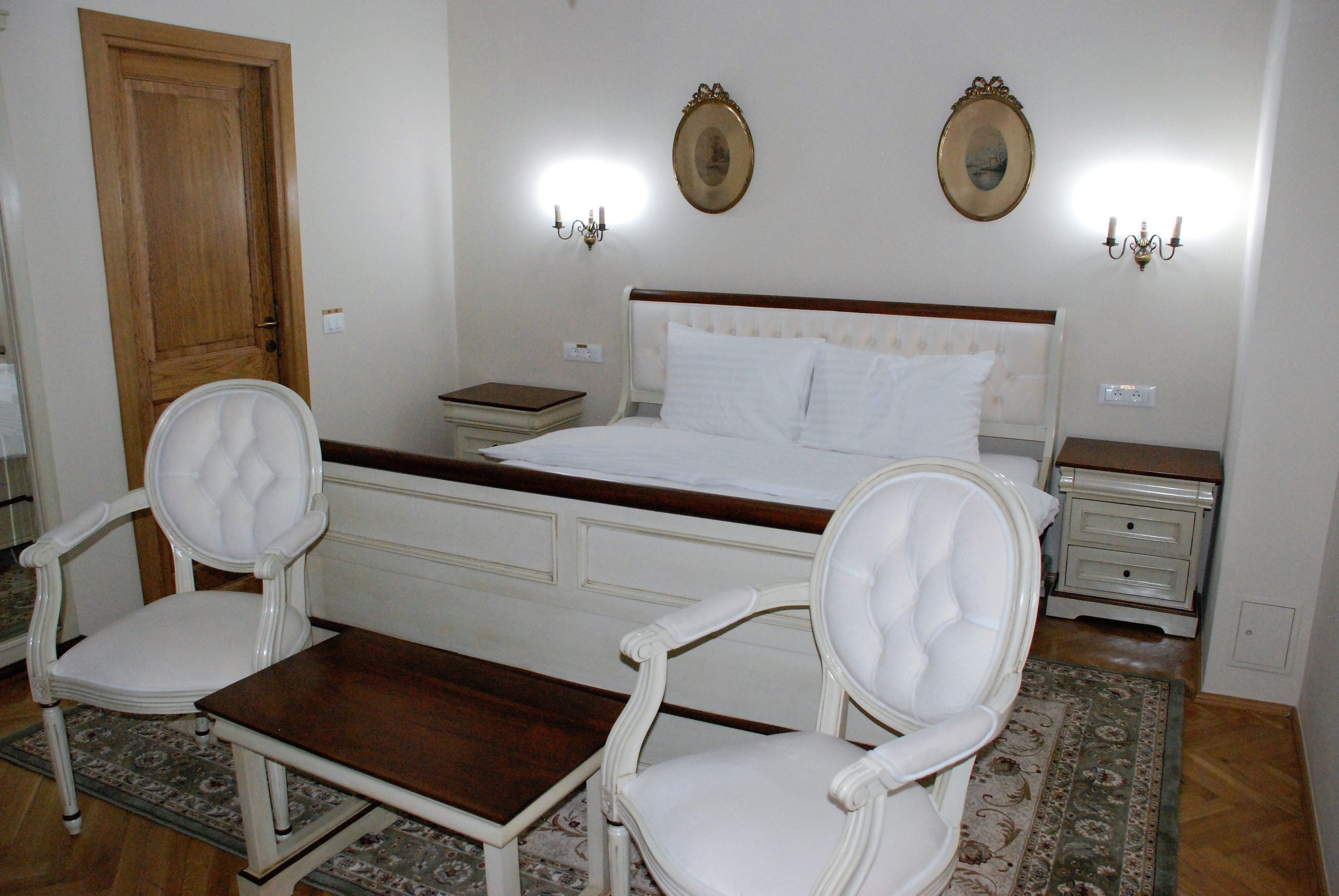 Un dormitorio con una cama, dos mesitas de noche, una mesa de madera, dos sillas tapizadas y una alfombra estampada.