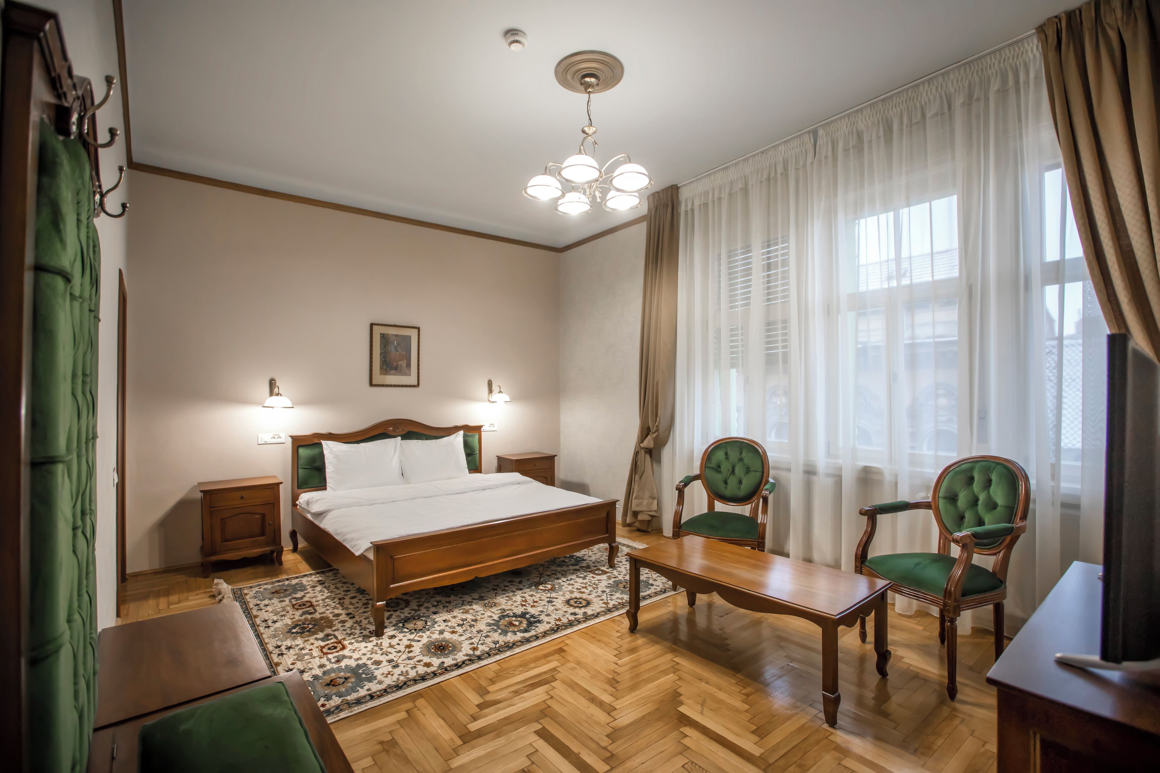 Habitació d'hotel amb un llit de fusta, un butac armchair verd, un taula de fusta i una finestra gran amb cortines.