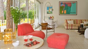 Uma sala de estar moderna com uma janela grande, um sofá vermelho, um pufe listrado e um espaço de jantar com mesa e cadeiras.