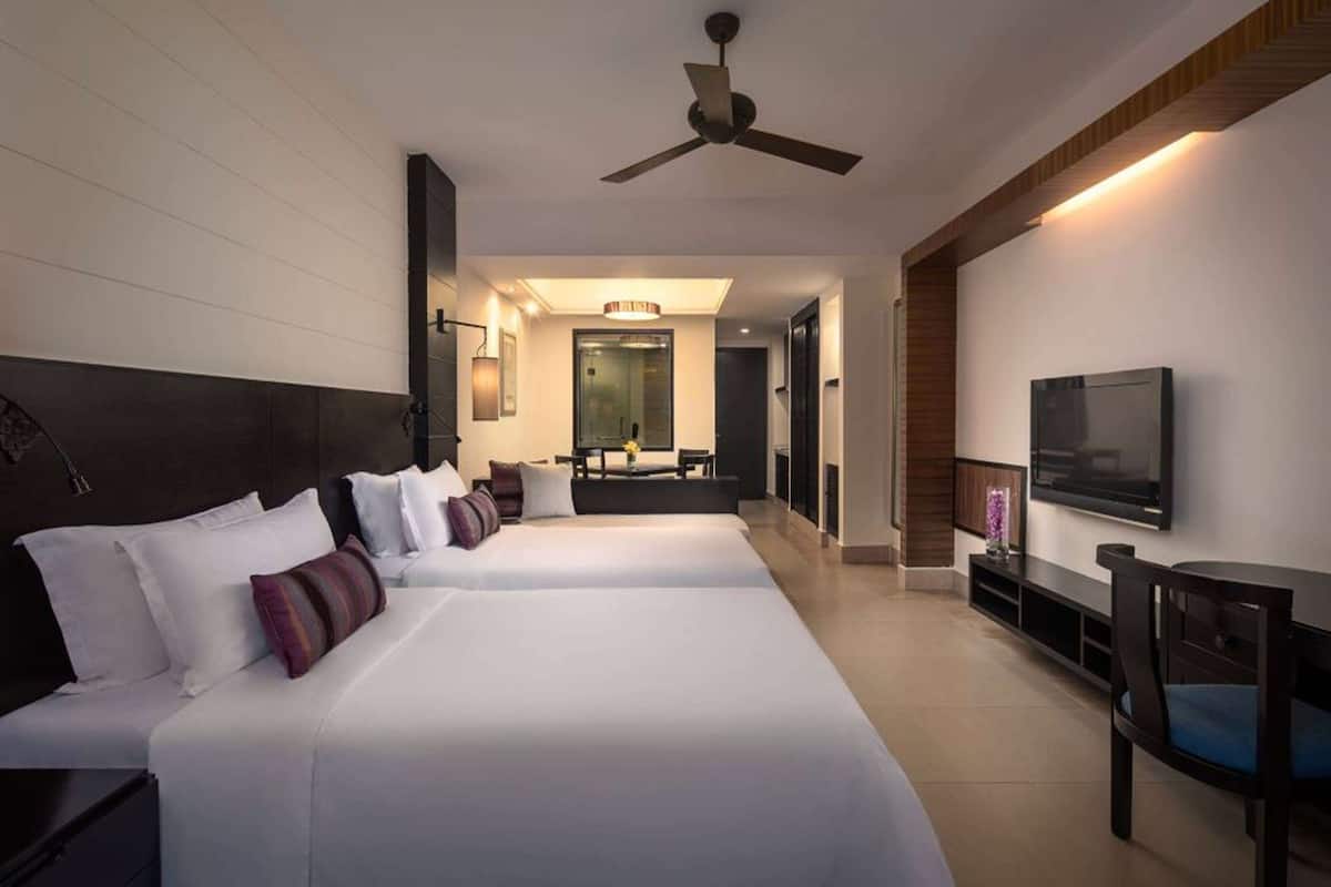 Habitación de hotel con dos camas, televisión, ventilador de techo y baño visible a través de una puerta abierta.