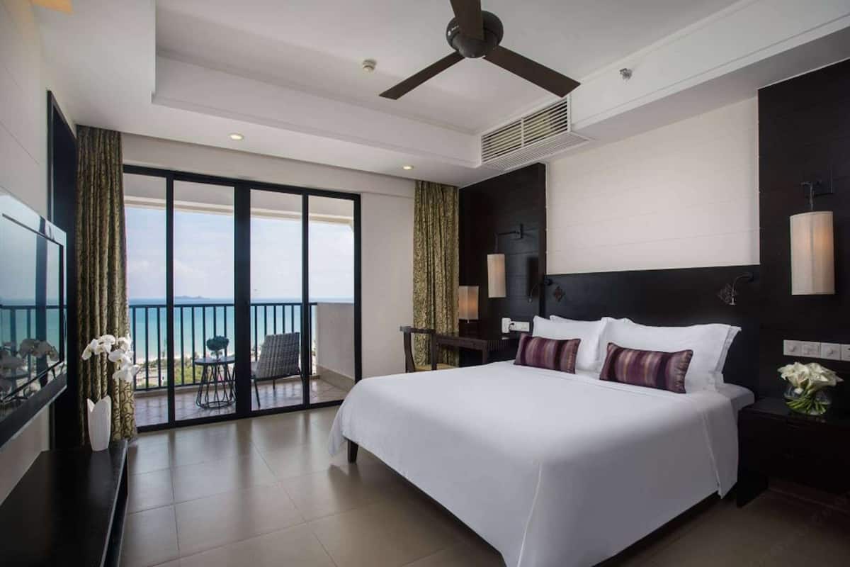Una habitación de hotel moderna con una cama grande, un ventilador de techo, un balcón con vistas al mar y un televisor a la izquierda.