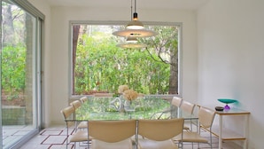 Een eetgedeelte met een glazen tafel, beige stoelen, een vaas met bloemen en uitzicht op groen door grote ramen.