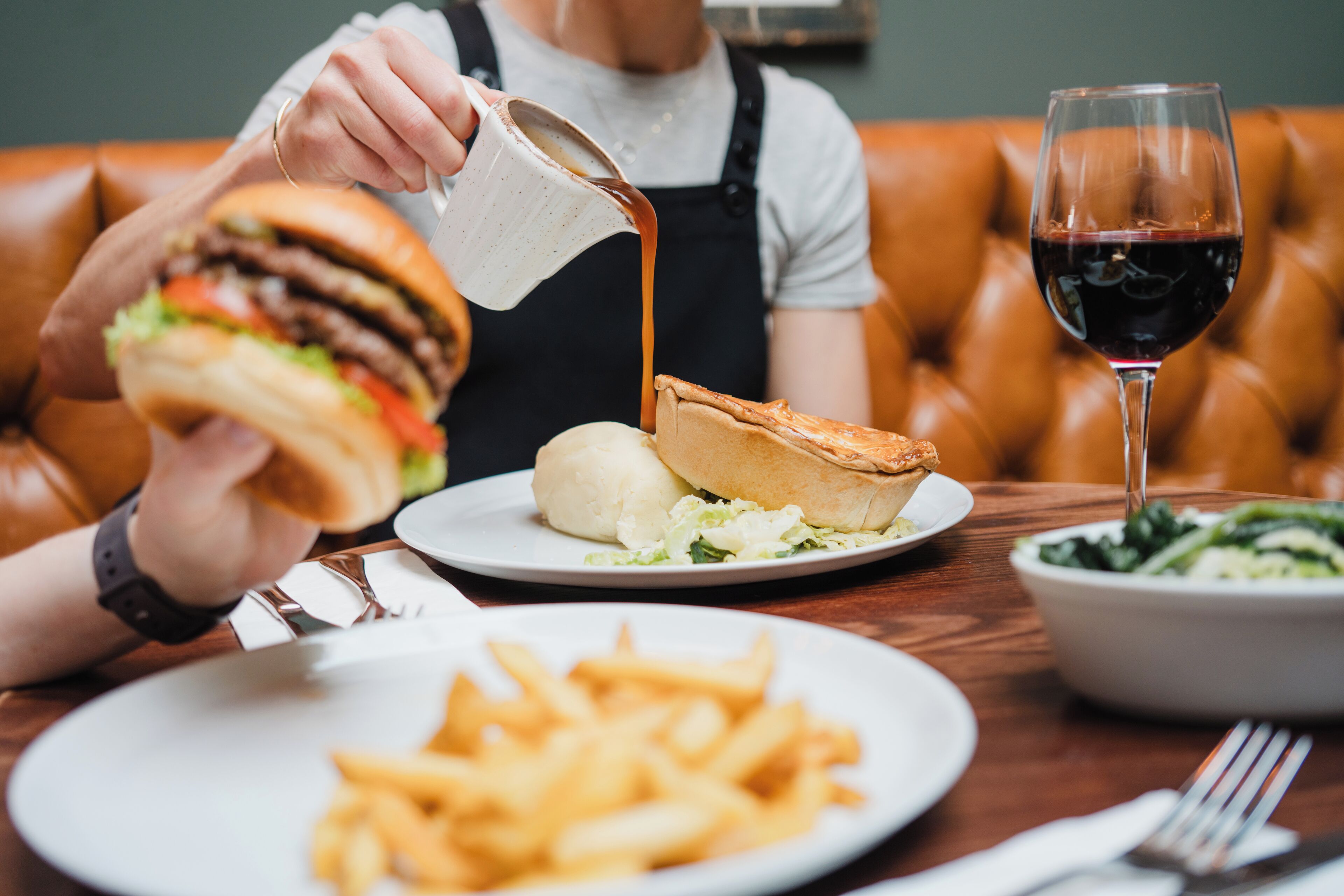 A dining table with a burger, a pie, a glass of red wine, and a bowl of greens.