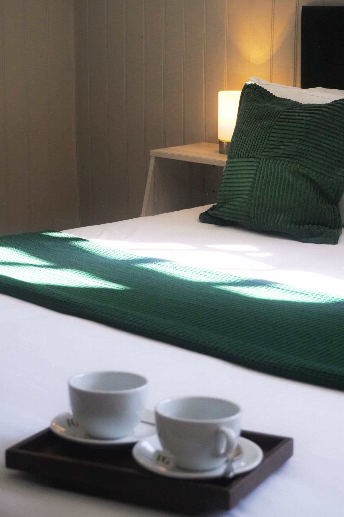 A neatly made bed with a green blanket, a pillow, and two cups on a tray.