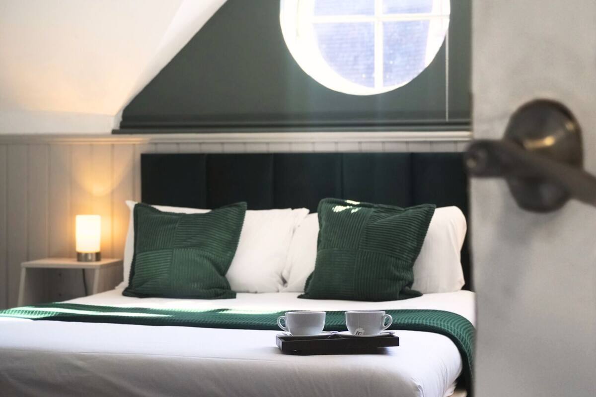 A neatly made bed with green pillows, a bedside table with a lamp, and a tray with cups and a teapot.