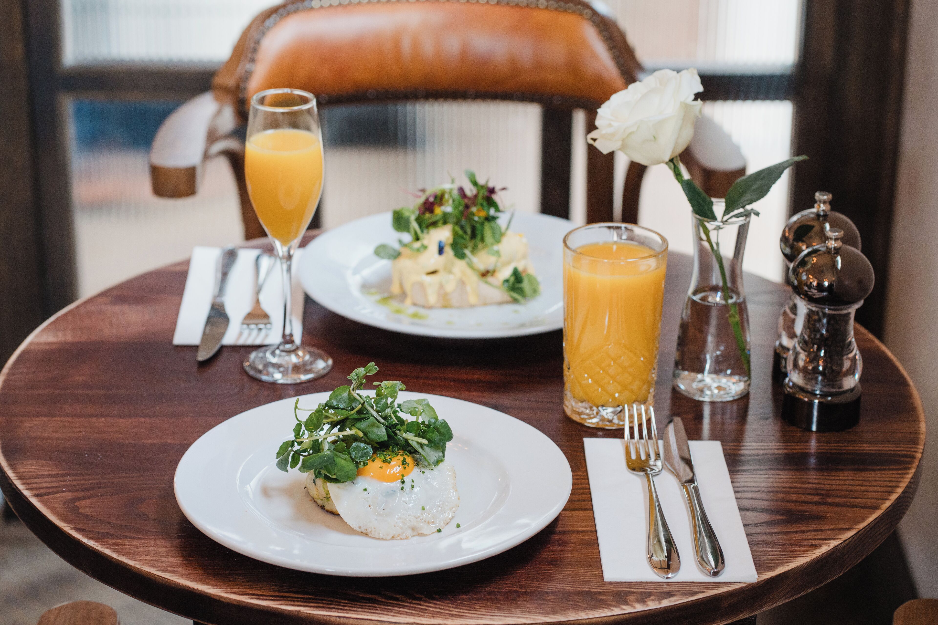 A dining table with a plate of food, a glass of orange juice, and a vase with a white rose.