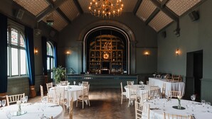 A spacious dining area with high ceilings, arched windows, and a central chandelier. Tables are set with white tablecloths and chairs.