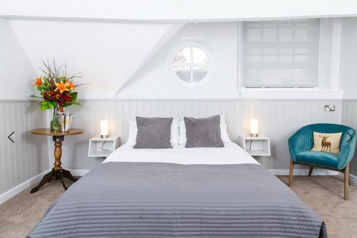 A bedroom with a large bed, two bedside tables, a round window, a chair, and a wall-mounted shelf.
