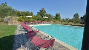Une piscine entourée de verdure et bordée d’un chemin de pierre, avec des chaises longues rouges et des parasols.