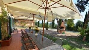 Une terrasse extérieure dotée d’une longue table et de chaises, protégée par de grands parasols et entourée de verdure.