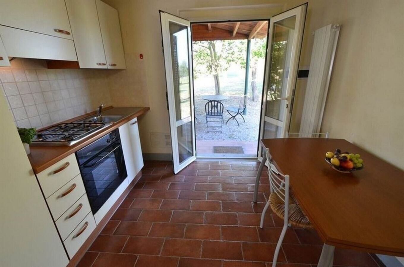 Une cuisine compacte équipée d’une cuisinière à gaz, d’un four et de meubles blancs. On y trouve une table à manger avec un panier de fruits, ainsi qu’une porte-fenêtre coulissante donnant sur un espace extérieur avec une table et des chaises de patio.