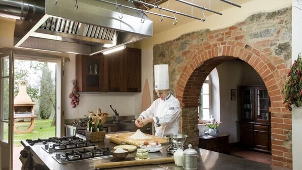 Un chef prépare des plats dans une cuisine dotée d’un grand îlot central, d’une arche en briques et offrant une vue sur l’extérieur.