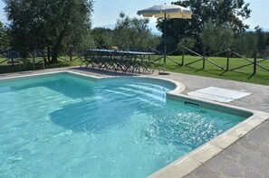 Ein rechteckiger Pool mit einer Steinterrasse, einem Metallzaun sowie einer Liege mit Sonnenschirm.