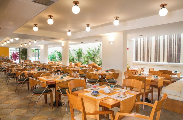 A spacious dining area with multiple tables and chairs, large windows, and indoor plants.