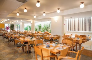 A spacious dining area with multiple tables and chairs, large windows, and indoor plants. - Flisvos Royal Tolo (Nafplio)