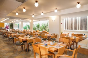Un comedor amplio con varias mesas y sillas, ventanas grandes y plantas de interior.