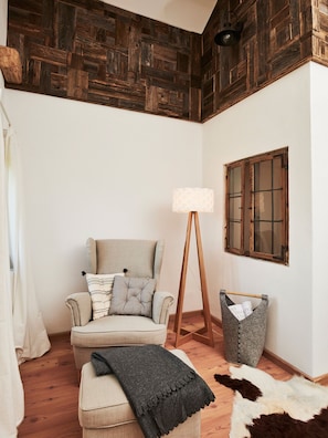 A cozy room with a wooden ceiling, a chair with cushions, a floor lamp, a window with wooden shutters, and a rug with a cowhide pattern.