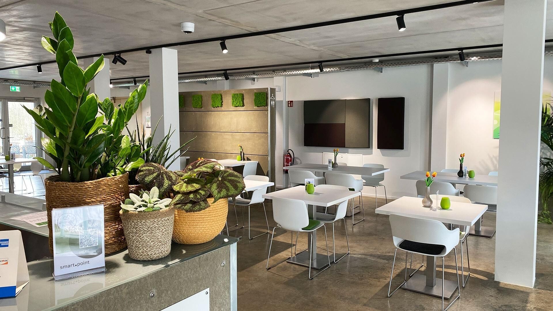 Un espacio de oficina moderno con mesas y sillas blancas, plantas en macetas y una pantalla grande en la pared.