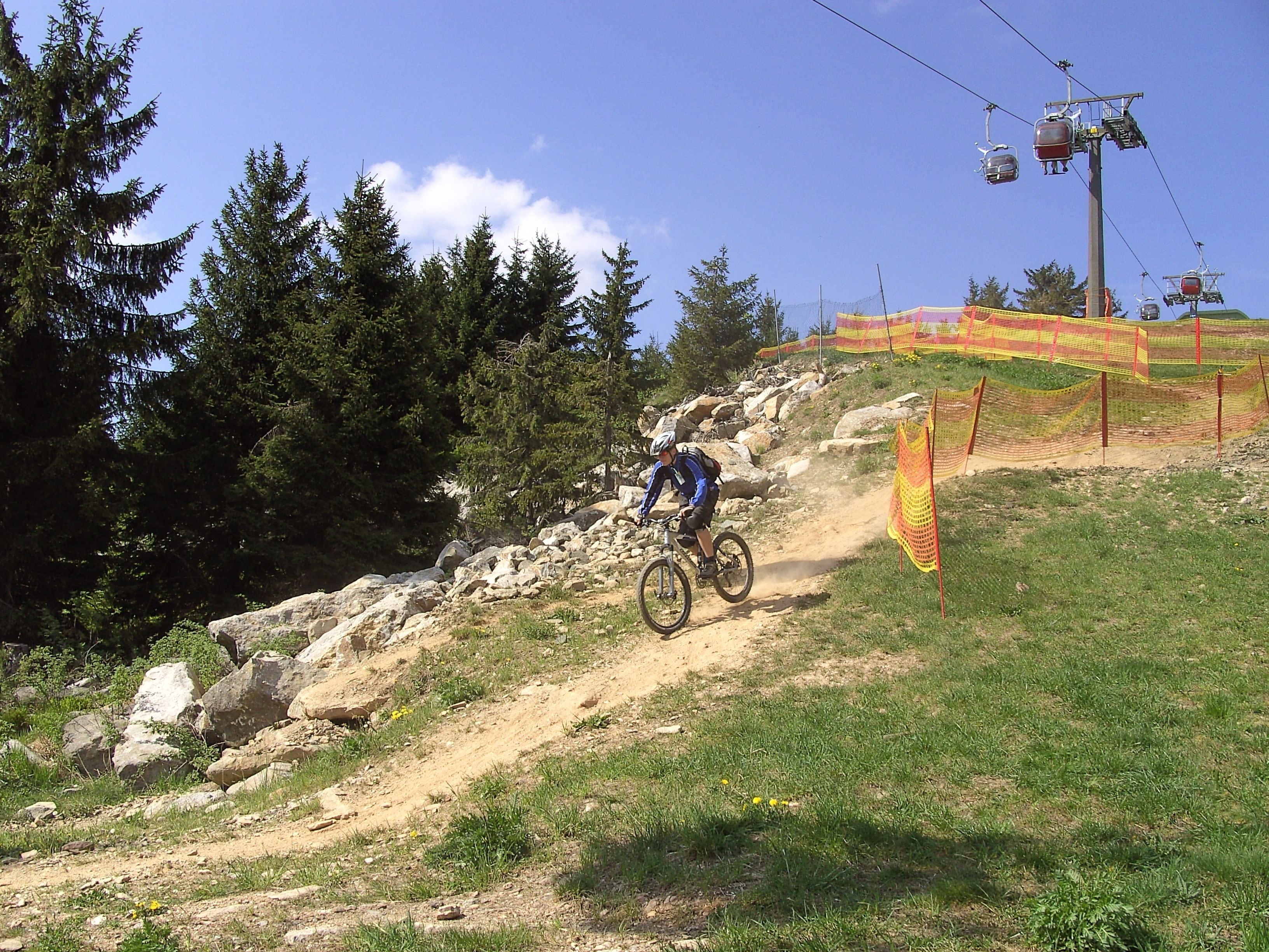 A mountain biker on a dirt trail with trees and a ski lift in the background.