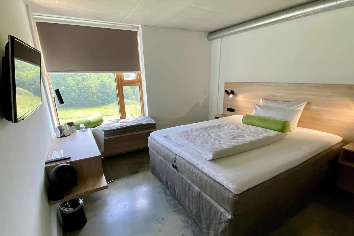 Un dormitorio con cama, mesitas de noche, escritorio, silla y ventana con vistas a zonas verdes.