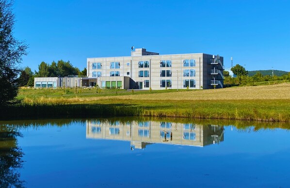 Un bâtiment moderne à plusieurs étages, doté de grandes fenêtres et dont le reflet se mire dans une étendue d’eau calme.