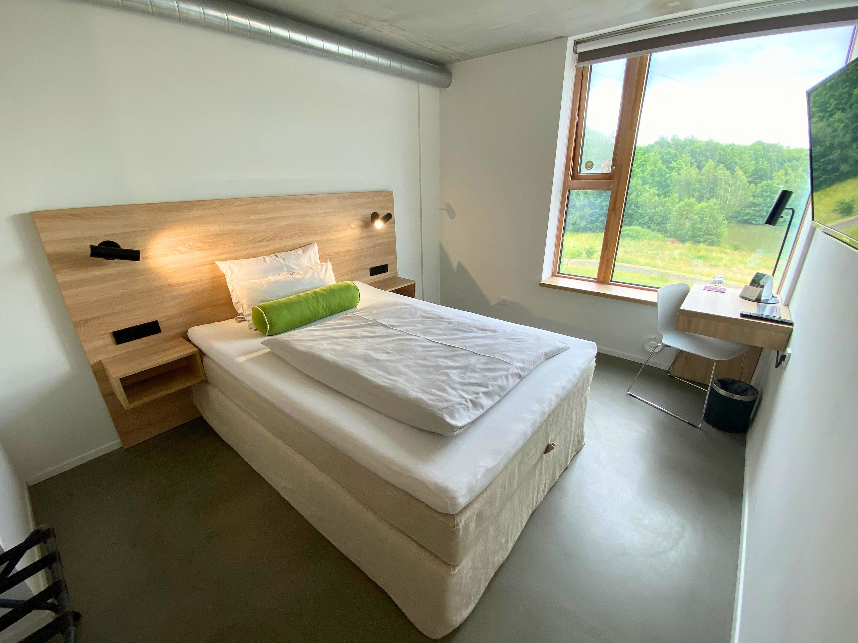 A bedroom with a wooden headboard, a bed with white linens, a green pillow, a desk with a chair, and a window with a view of greenery.