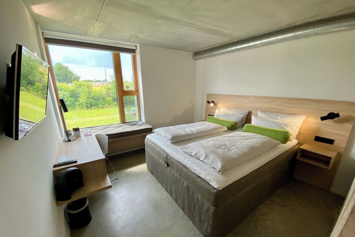 Un dormitorio con cama, escritorio, silla, televisor y ventana con vistas a la vegetación.