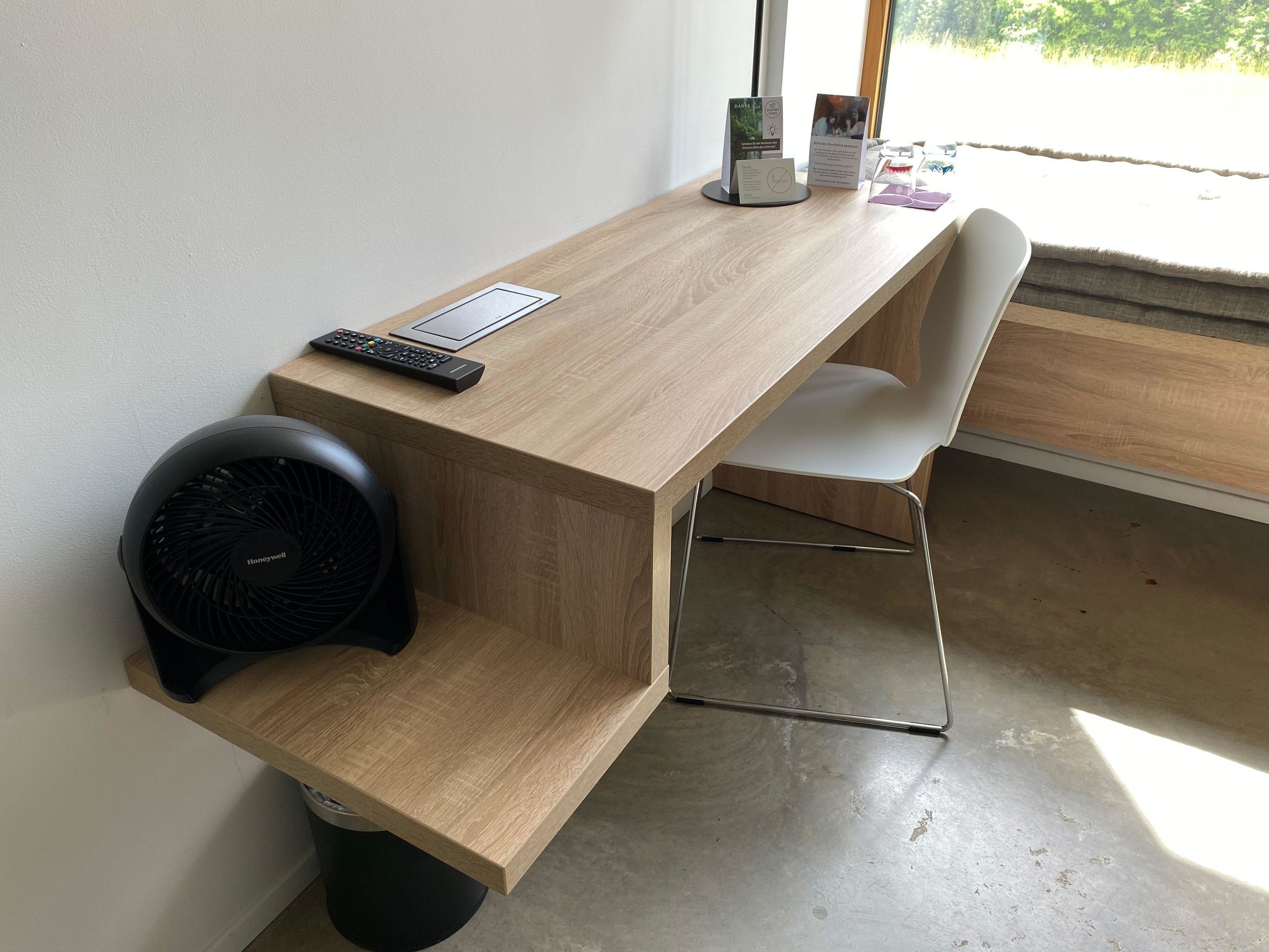 A wooden desk with a remote control, a fan, and a chair.
