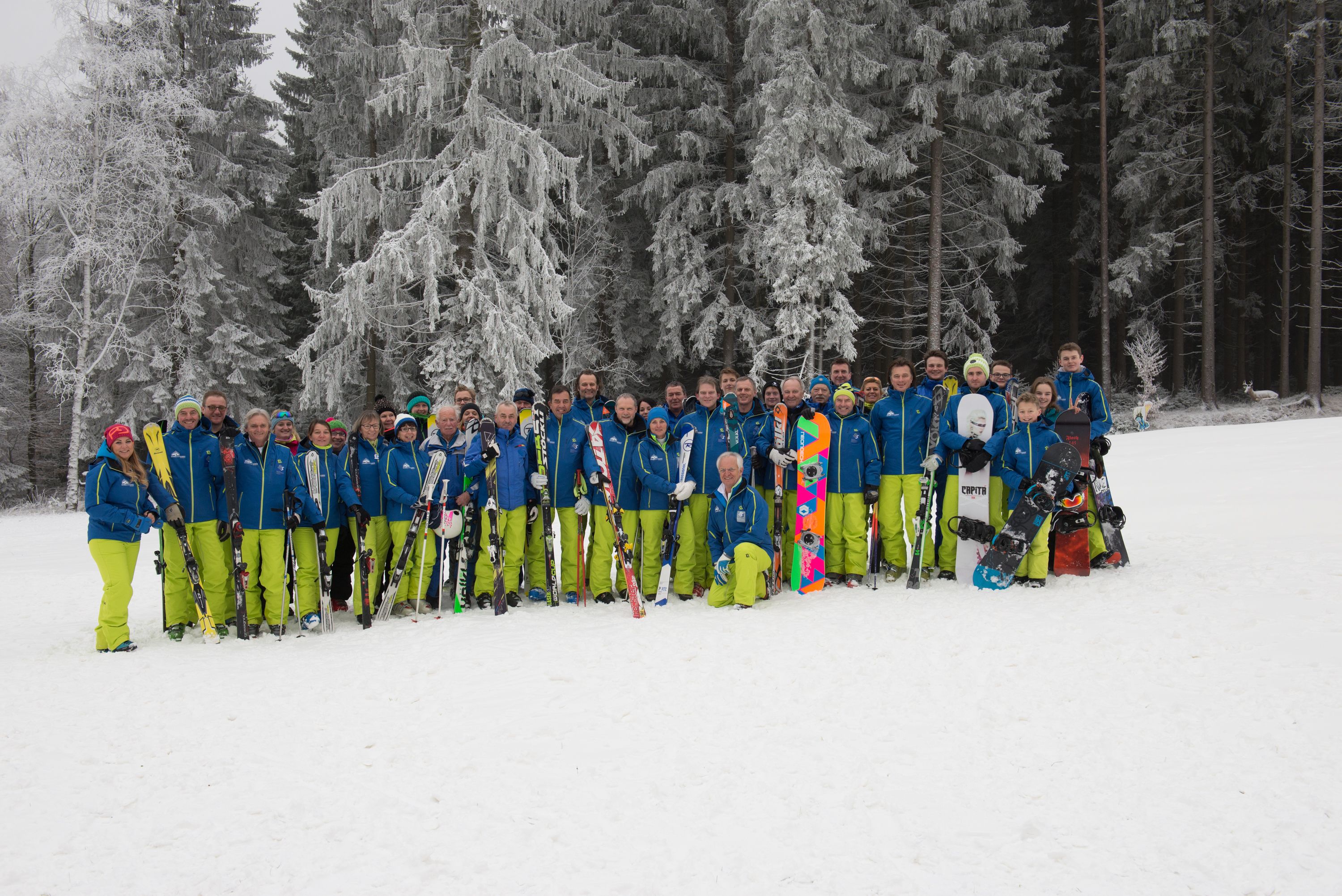A group of people in blue jackets and green pants standing in a snowy area with skis and snowboards.
