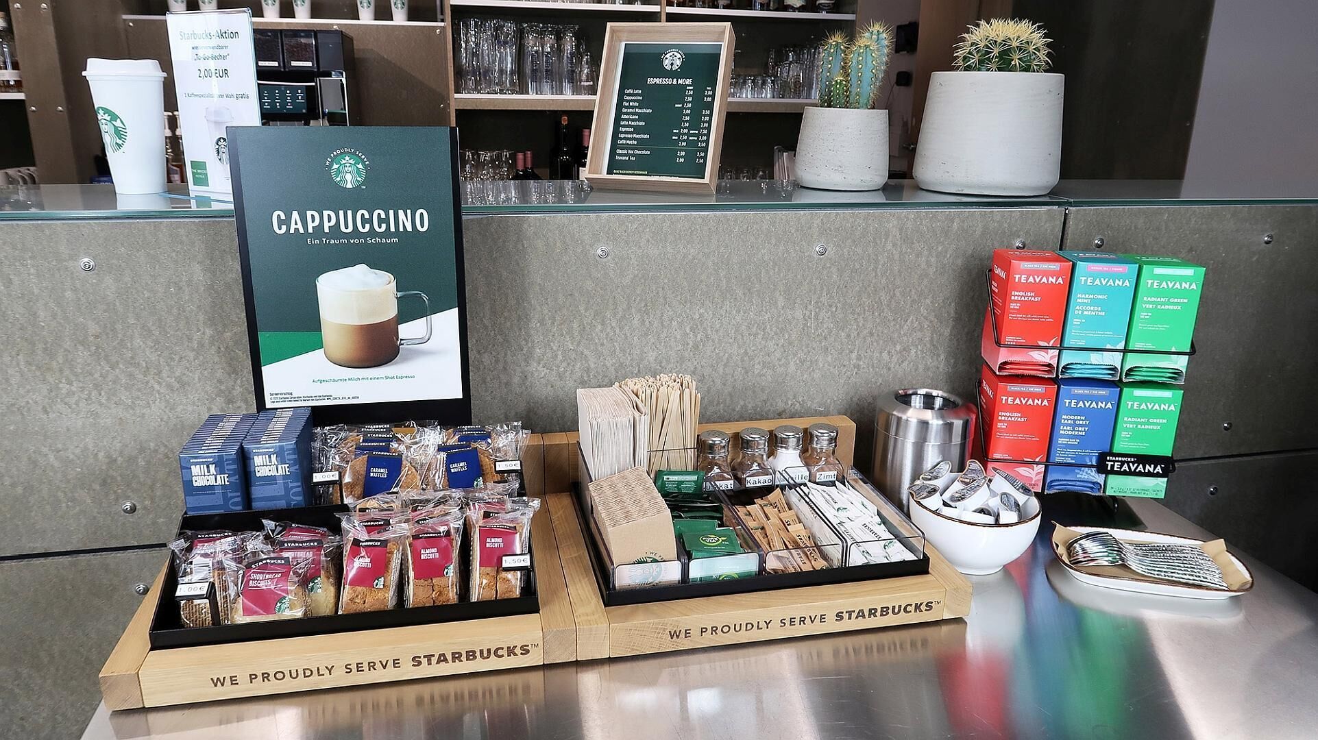 Eine Starbucks-Auslage mit einem Cappuccino-Schild, verschiedenen Kaffeeboxen und Teebehältern sowie einer Platte mit Cantuccini.