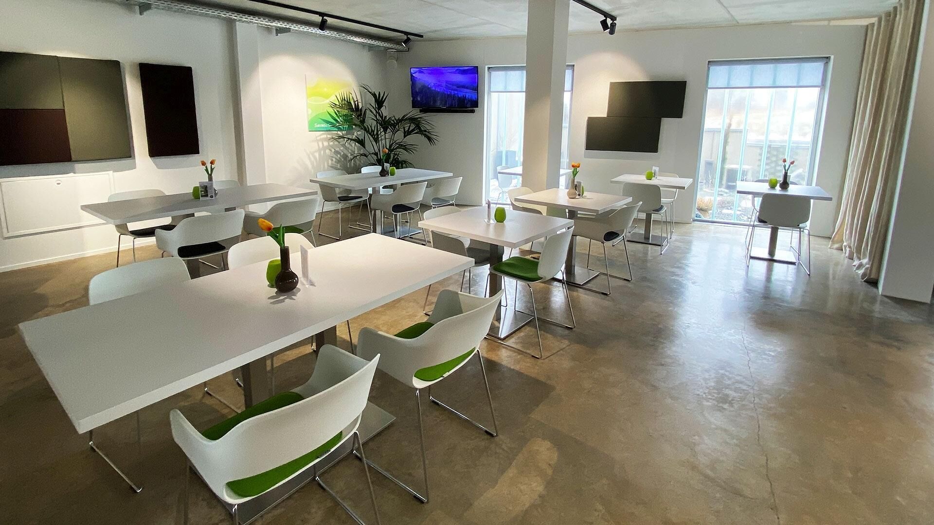 A modern office space with multiple white tables and green chairs, large windows, and a flat-screen TV on the wall.