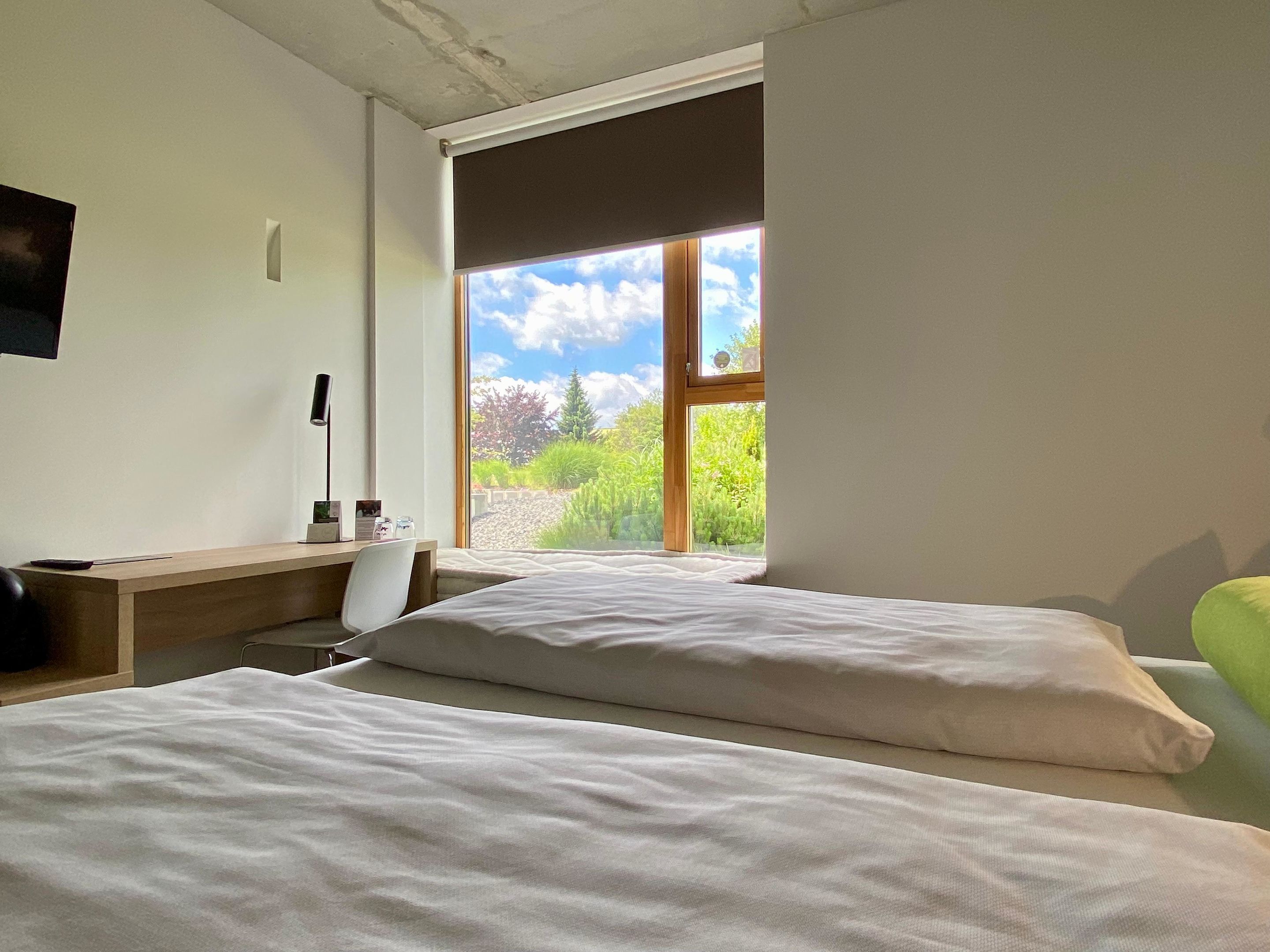Un dormitorio con dos camas, un escritorio y una silla. Se ve una ventana con vistas a la vegetación y un cielo despejado.