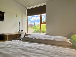 A bedroom with two beds, a desk, and a chair. There is a window with a view of greenery and a clear sky. - Avaneo Hotel Marktredwitz (Marktredwitz)