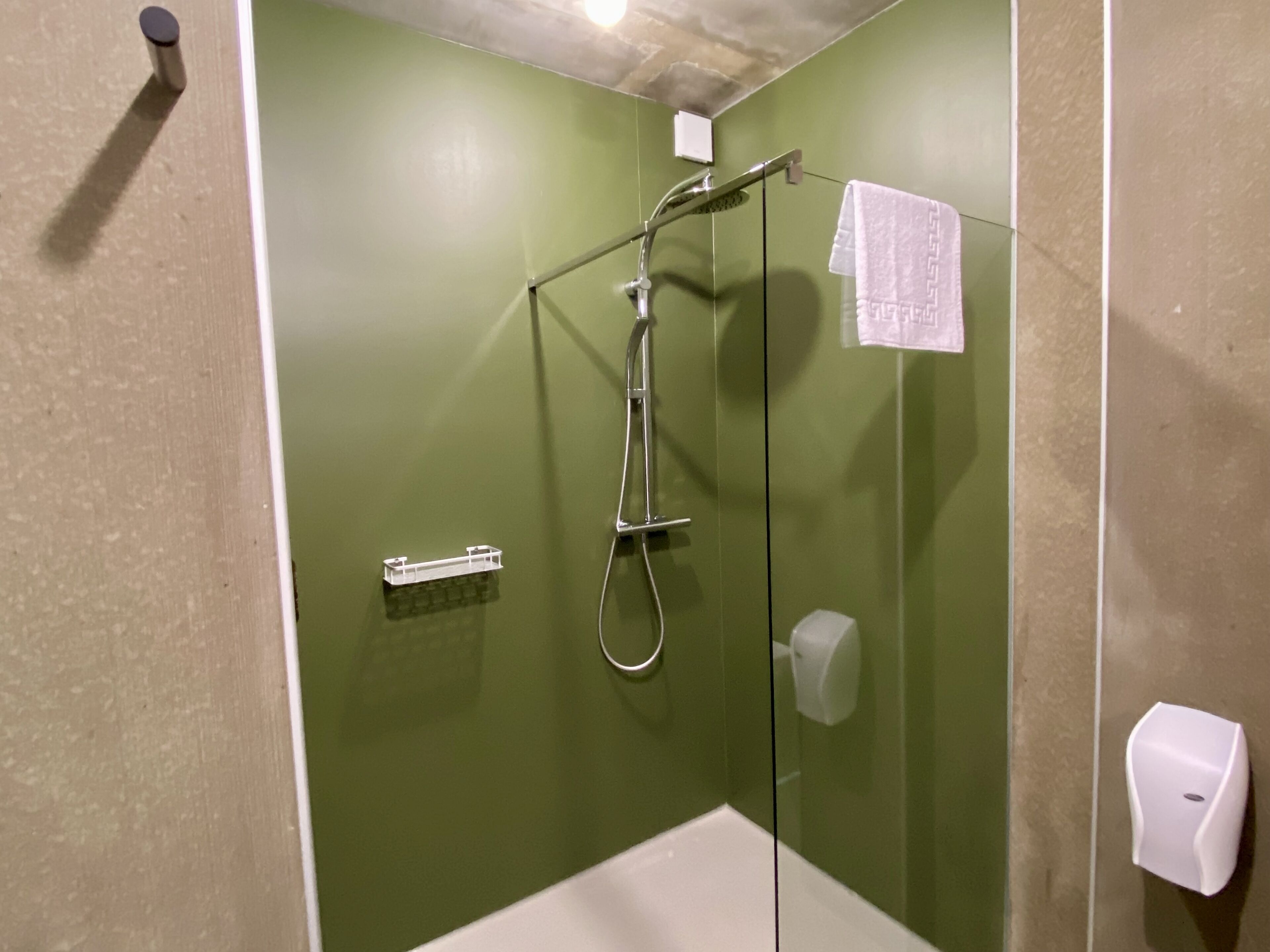 Cabina de ducha con mampara de vidrio, cabezal de ducha manual, dispensador de toallas y dispensador de jabón.