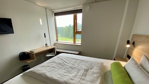 Una habitación de hotel con cama, escritorio, silla, ventana con vista a follaje y lámpara fijada a la pared.