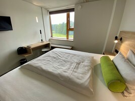 Quarto de hotel com cama, escrivaninha, cadeira, janela com vista para vegetação e luminária na parede.