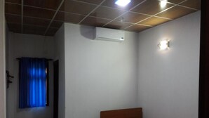 A small, single-bed room with a wooden headboard, a window with blue curtains, a wall-mounted air conditioner, and a small wall-mounted light fixture. - Ulpatha Nature Resort (Kobeigane)