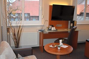 A hotel room with a wooden desk, a flat-screen TV, a round table with a plate of fruit, a vase with flowers, and a view of buildings through the window.