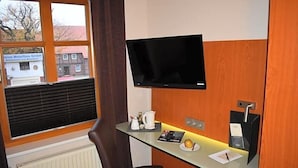 A hotel room with a large window, a flat-screen TV mounted on the wall, a desk with a kettle, a plate with a fruit, and a chair.