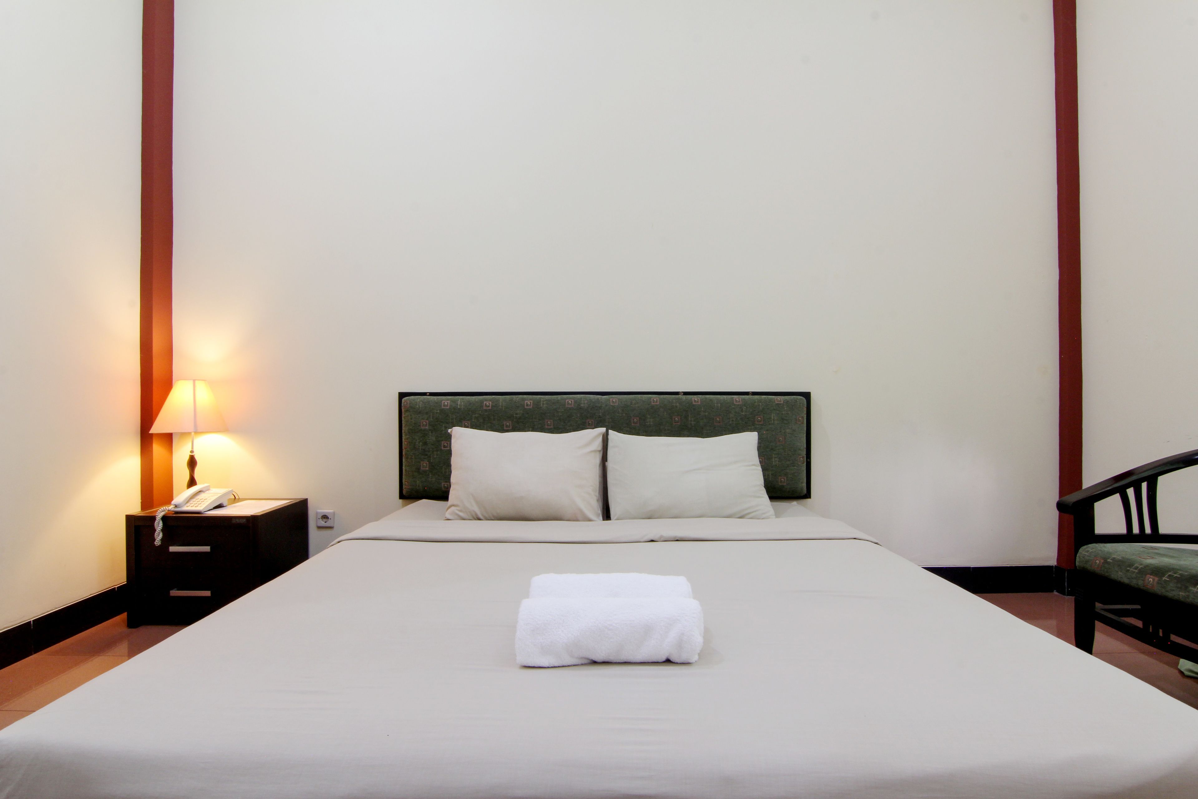 Una cama bien hecha con una toalla blanca doblada, una lámpara de noche y un teléfono en la mesita.