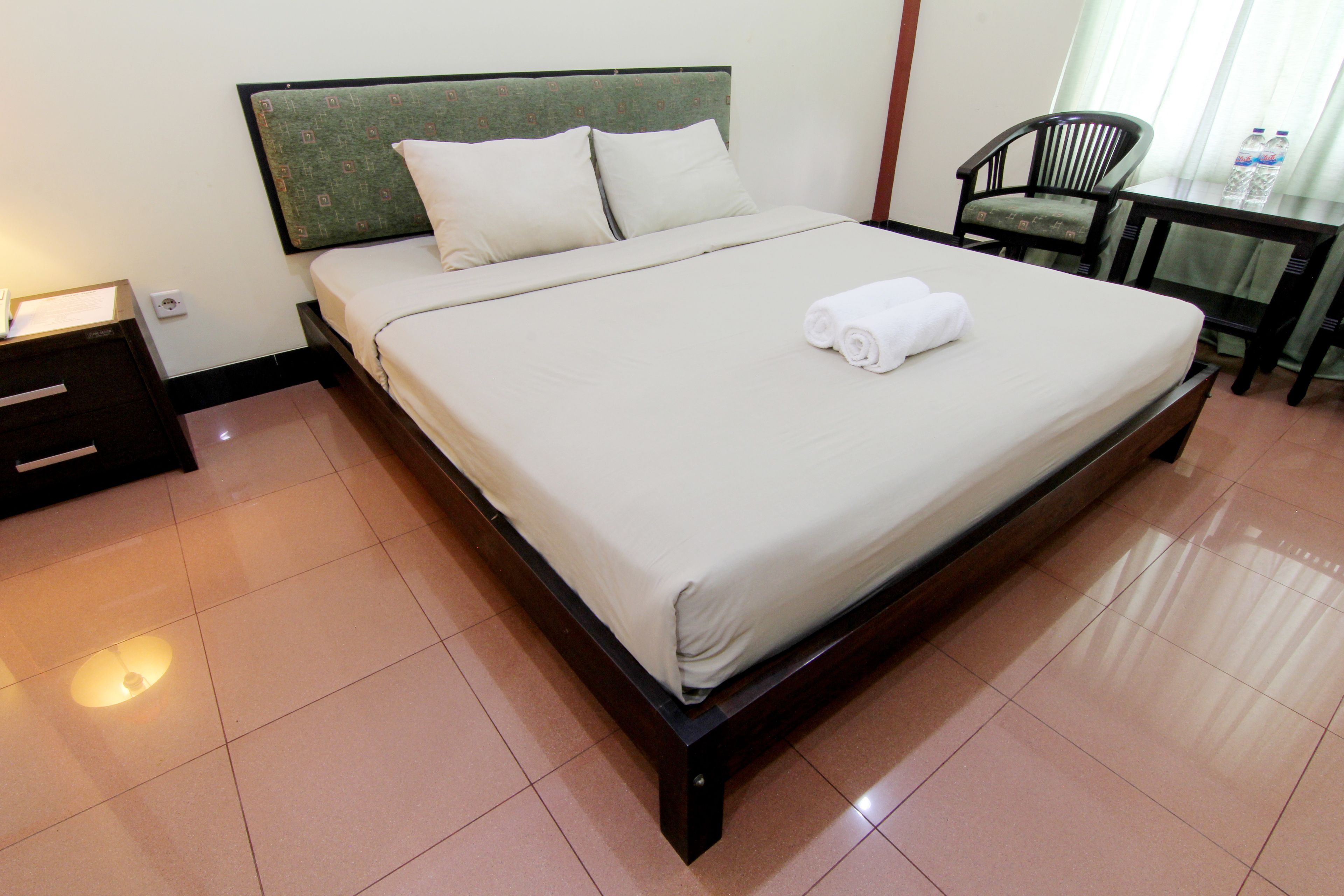 A neatly made bed with white linens and rolled towels, a wooden headboard, and a small table with chairs in a hotel room.