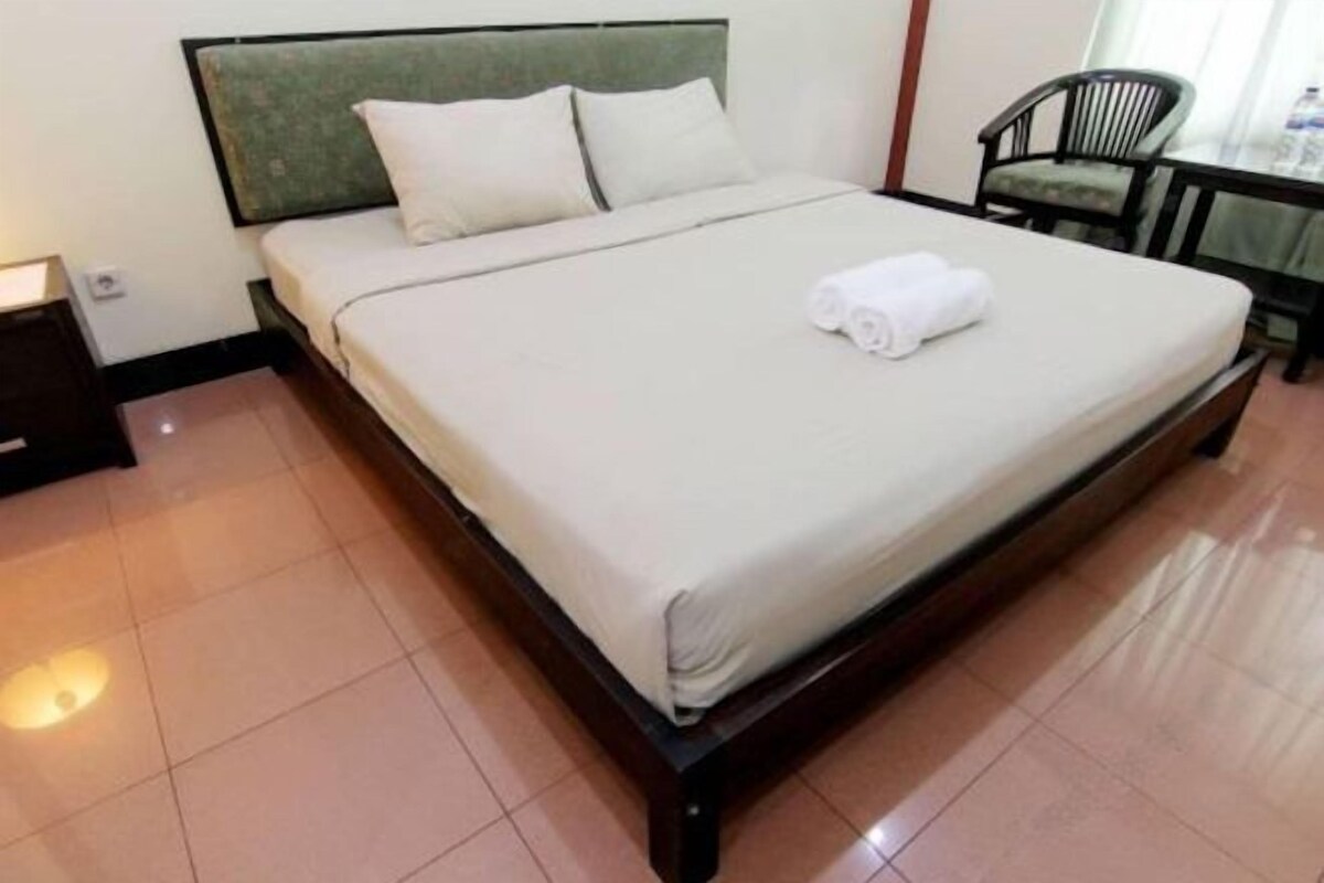 A neatly made bed with a green headboard and white linens, a small table with a chair, and a tiled floor.