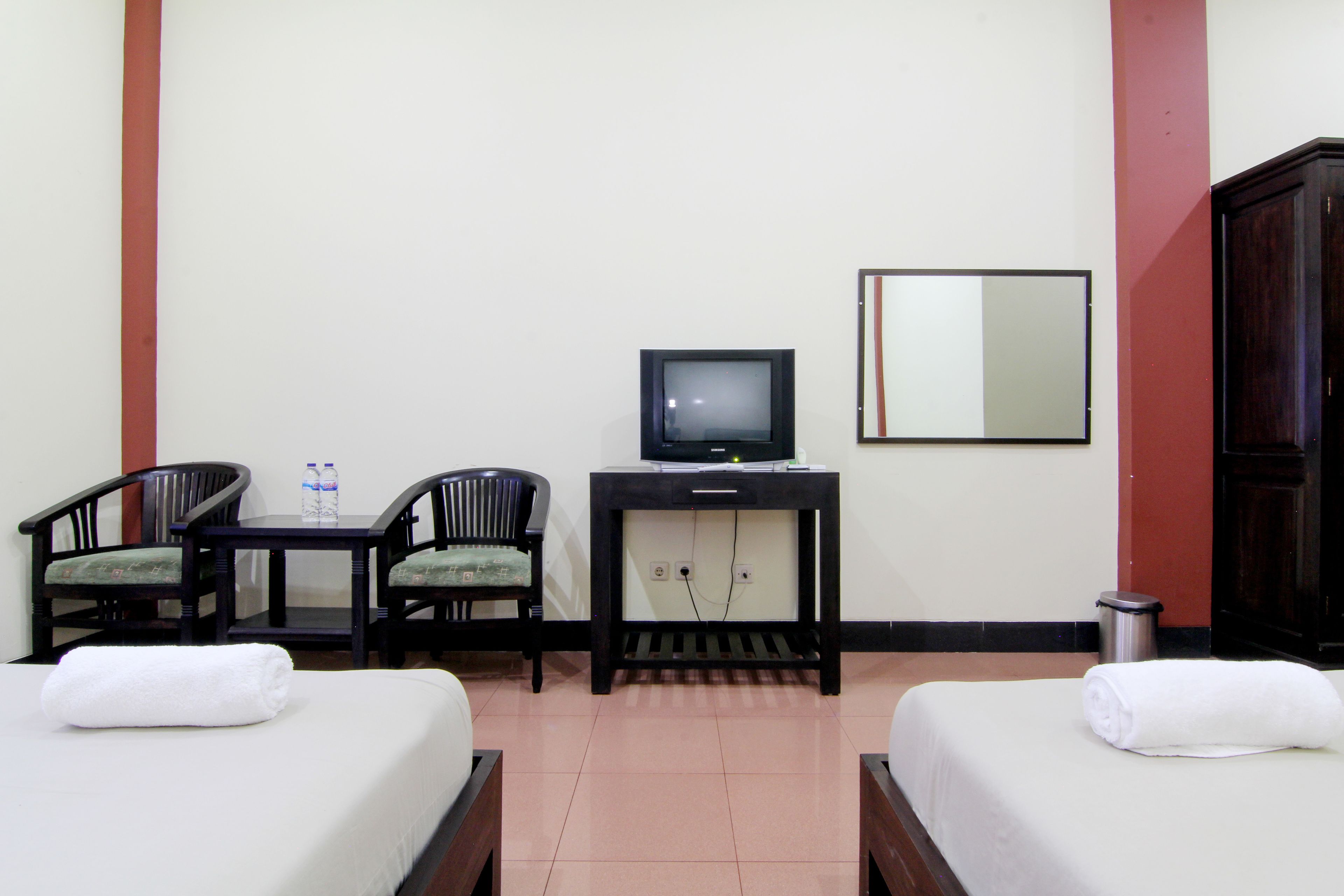 A hotel room with two beds, a desk with a television, two chairs, and a trash bin.